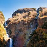 岩壁を流れ落ちる称名滝が陽光に照らされ、周囲の紅葉とともに輝いている風景。