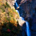 岩壁を流れ落ちる称名滝が陽光に照らされ、周囲の紅葉とともに輝いている風景。