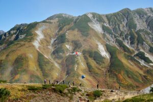 雄大な立山の山肌を背景に、青い荷を吊り下げたヘリコプターが飛行し、下には登山者が並んでいる