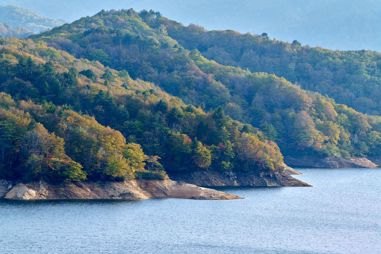 有峰湖の湖岸に迫る紅葉した山の斜面。赤や橙、黄色に染まった木々が湖面に沿って広がる風景