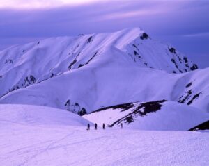 紫がかった夕暮れの空の下、白銀に覆われた奥大日岳の斜面を背景に、雪原を歩く登山者たちのシルエット