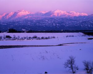 富山市内の雪原越しに、夕暮れの光で赤紫に染まる立山連峰を望む冬景色