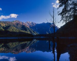 秋の大正池から望む穂高連峰。紅葉した岩肌と山影が湖面に映る風景写真。