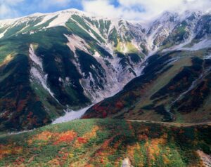 雄山の白い岩肌と山頂付近の初雪を背景に、山腹に赤や黄色の紅葉が広がる立山の秋景色